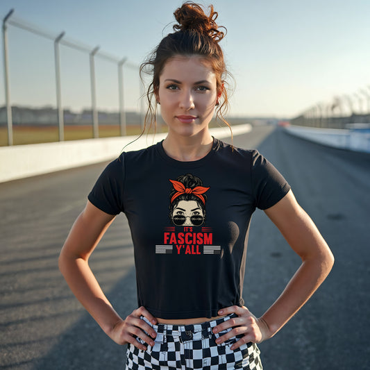 Woman standing confidently on a racetrack wearing a black crop top that says “It’s Fascism Y’all,” featuring a stylized face with barbed wire sunglasses and a red bandana, parodying “It’s Raceday Y’all.”
